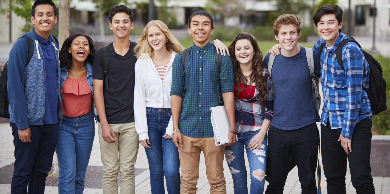 Eight students stand one beside the other and smiling. A student in the center holds a binder in his hand.